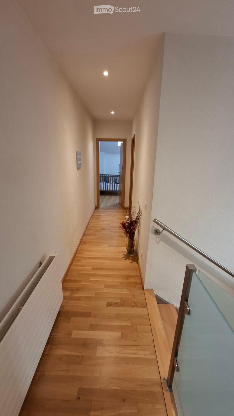 A narrow hallway with wooden flooring, white walls, and a metal handrail on the right side. A vase with dried flowers is placed on the floor near the railing, and a door at the end leads to another room.