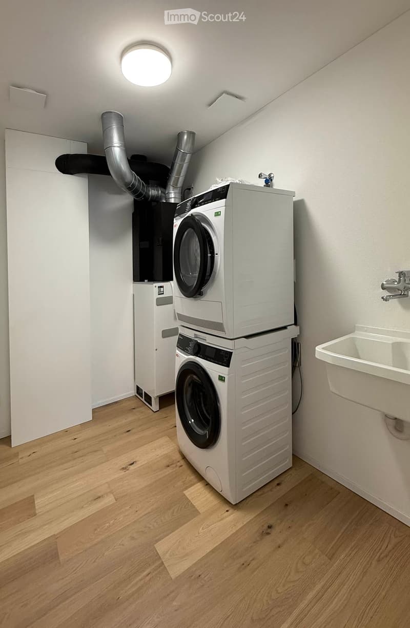 A small, well-lit room with wooden flooring, featuring a stacked washing machine and dryer unit, a sink, and visible ventilation pipes.