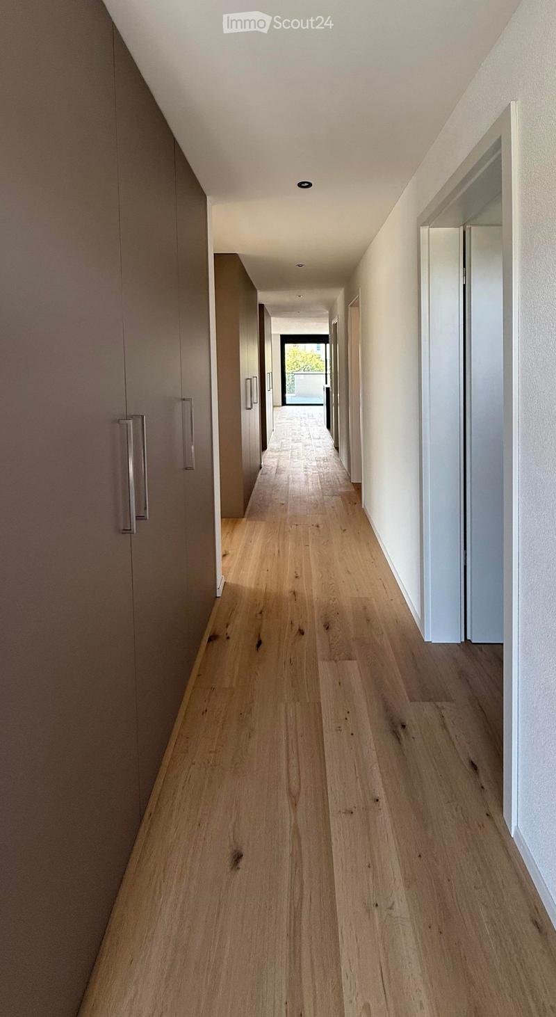 Modern hallway with light wooden flooring, white walls, and built-in storage units with silver handles. The corridor leads to an open area with natural light.