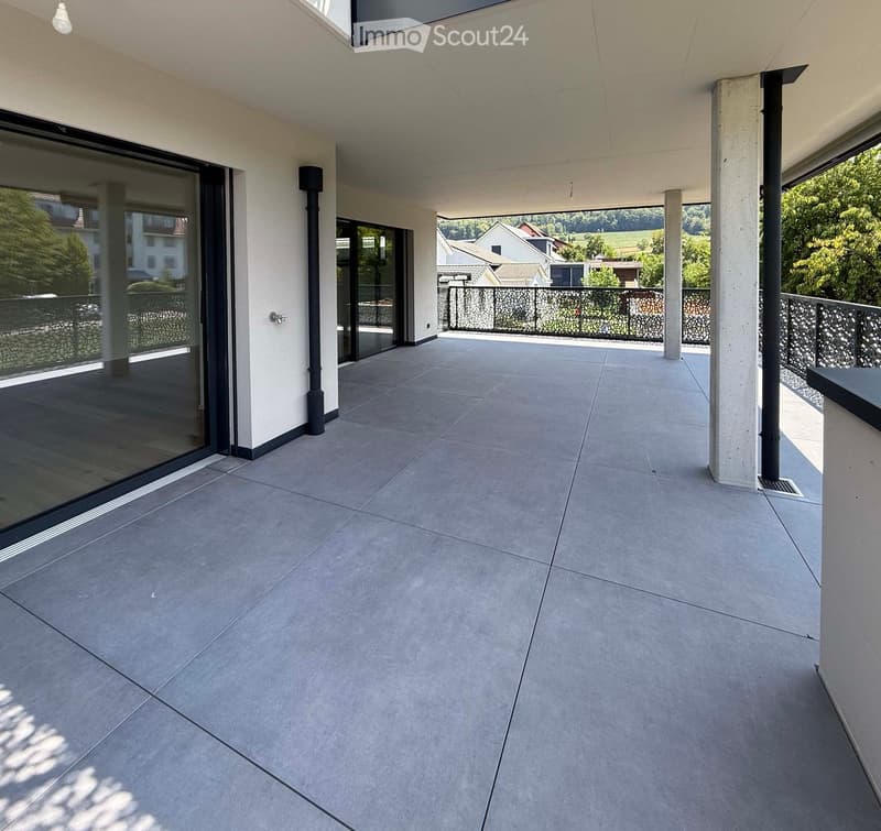 Modern balcony with large sliding glass doors, grey tiled floor, white walls, and a view of the surrounding landscape.