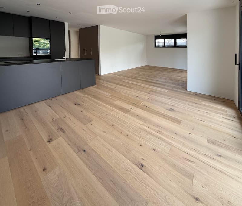 Empty room with light wooden flooring, white walls, and a grey kitchen counter with cabinets visible in the background. No furniture or personal items are present.