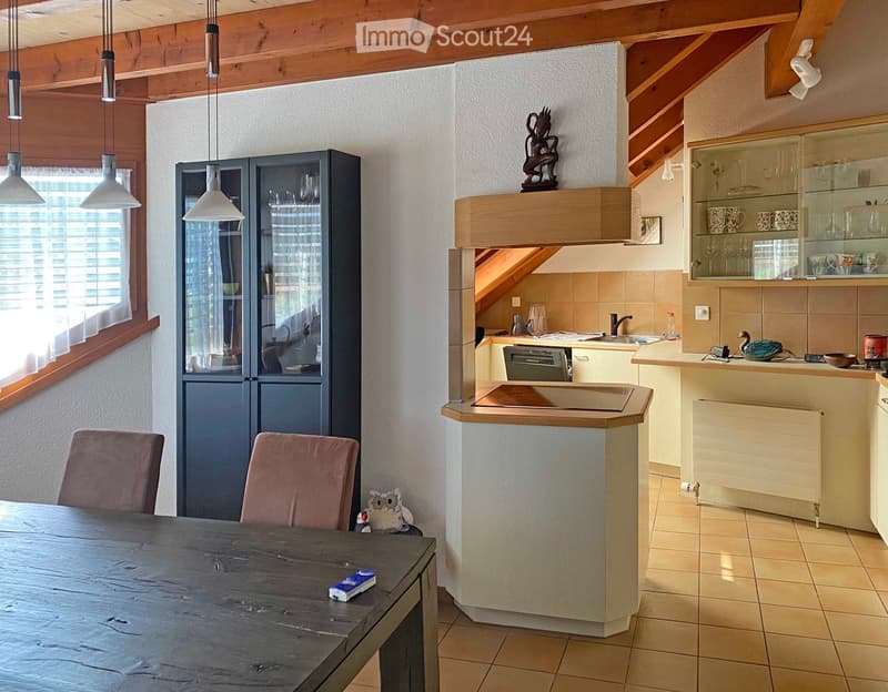 The image shows a cozy, well-lit kitchen and dining area with a wooden ceiling and exposed beams. The kitchen features a countertop, sink, dishwasher, and various appliances. A dining table with chairs is positioned near a large window with blinds. Shelve