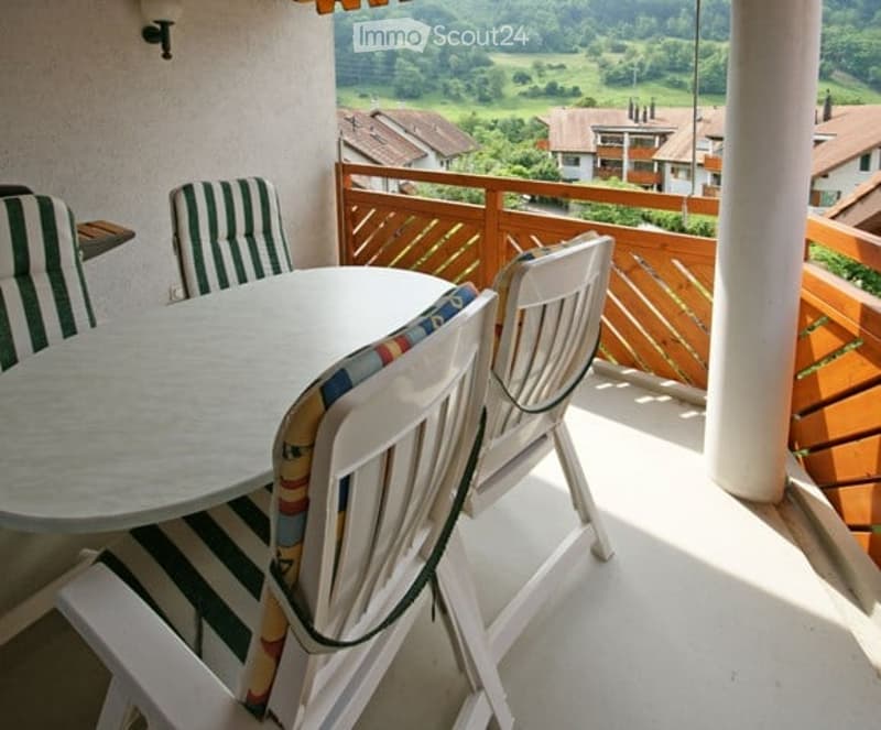 Balcony with a round table and four chairs, overlooking a scenic valley with houses and greenery.