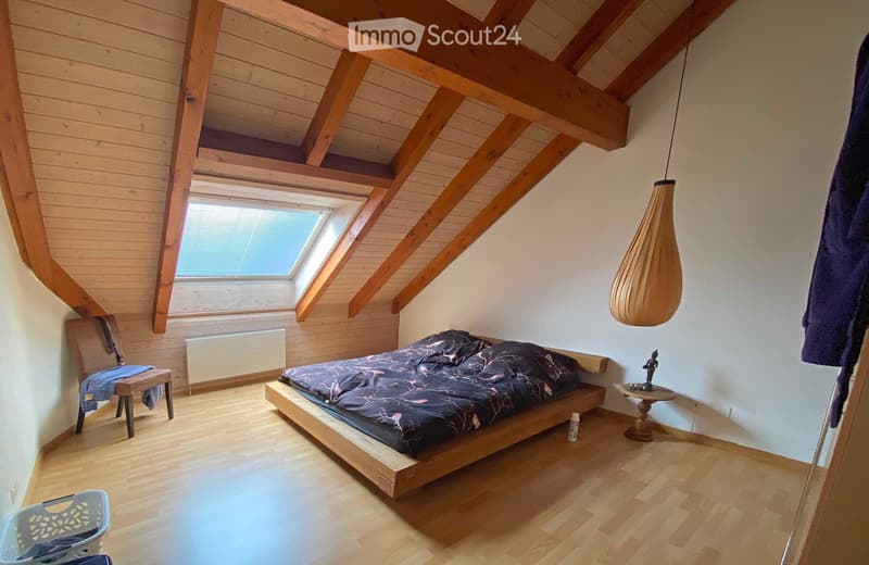 Cozy attic bedroom with wooden beams, skylight window, bed with dark bedding, small chair, side table, and hanging lamp.