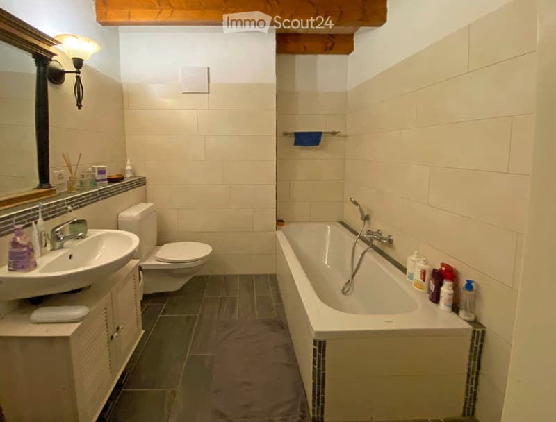 Bathroom with a sink, toilet, bathtub, mirror, and cabinet. The walls are tiled, and the floor is covered with a grey mat.