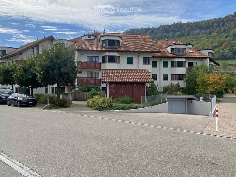 A large, modern two-story house with a red-tiled roof, white walls, multiple windows, and balconies. The house is surrounded by greenery and has a driveway with parked cars.