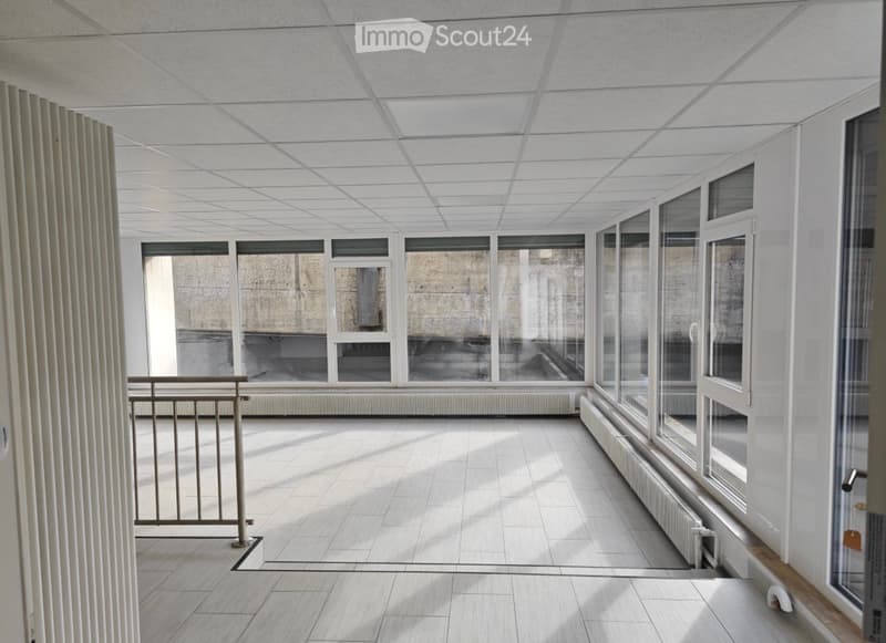 Empty room with white walls, tiled floor, large windows, and a metal railing, illuminated by natural light.