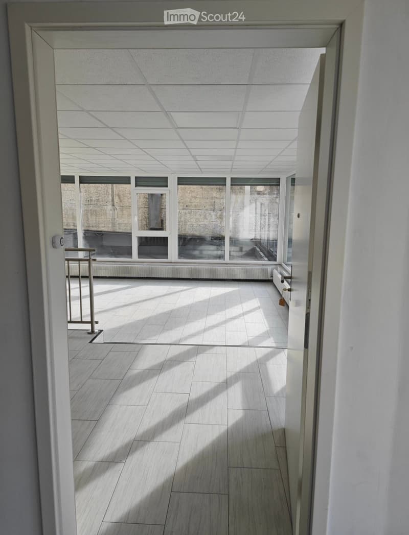 Bright, open corridor with white tiled floor and walls, large windows allowing natural light, and a metal railing on one side.