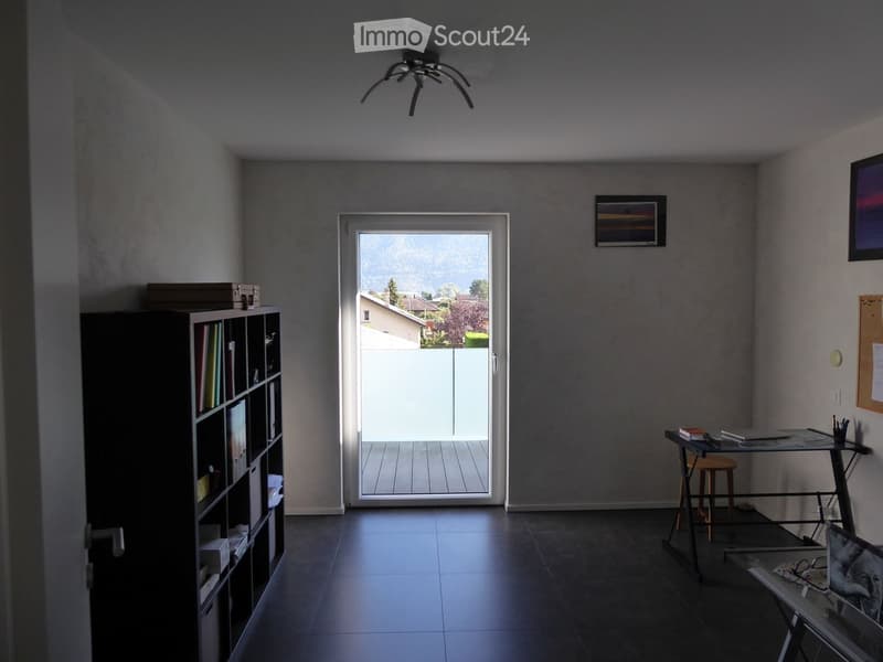 Bright room with dark tiled floor, large sliding glass door leading to a balcony with a scenic mountain view, bookshelf, small table with chair, and picture frame on wall.