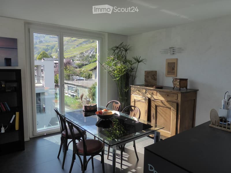 Bright dining area with a glass table, four chairs, a bowl of fruit, a tall plant, and a wooden cabinet. Large windows offer a scenic view of the valley.