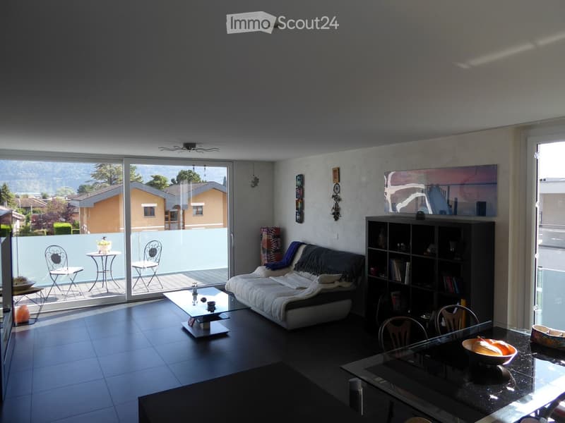 Modern living room with a sofa, glass coffee table, dining table, chairs, bookshelf, and a view of the outdoor area through large windows.