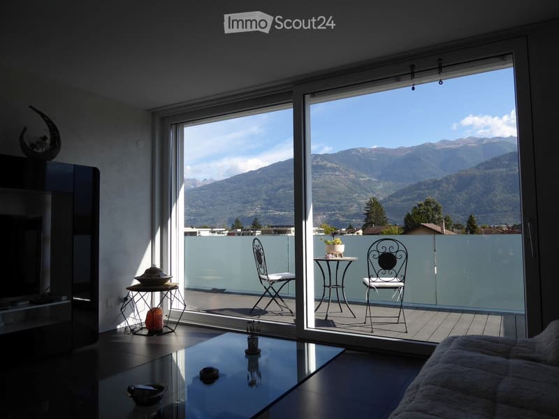 Modern living room with large glass doors opening to a balcony, offering a scenic mountain view. The room features a sofa, coffee table, and decorative items, while the balcony includes a small table and chairs.