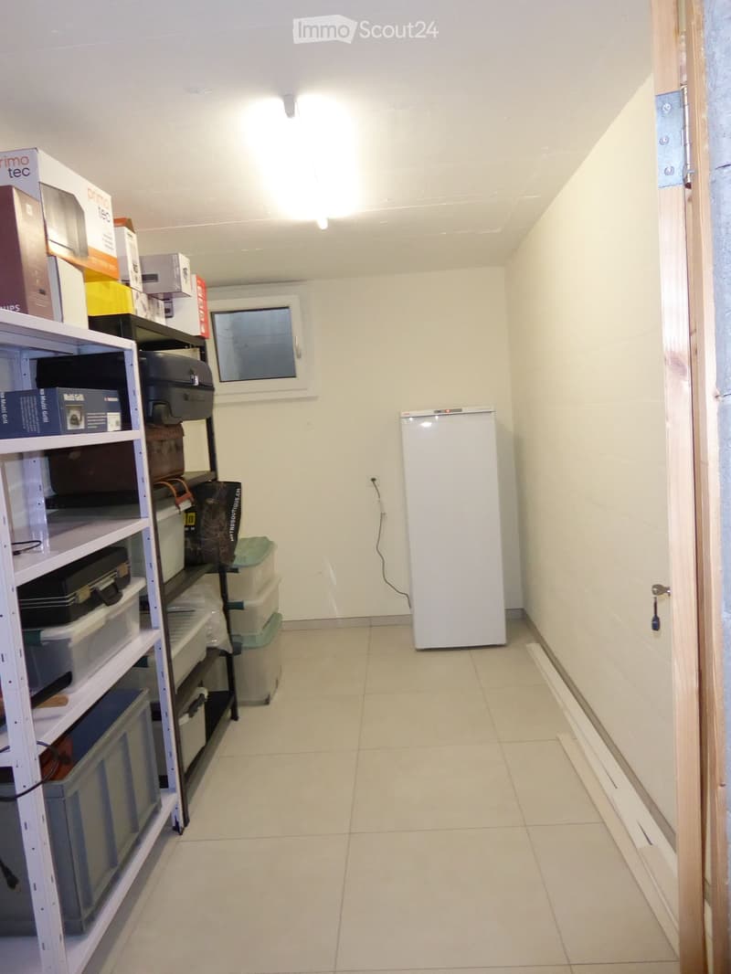 A small, well-lit storage room with a white tiled floor, white walls, and a shelf filled with various storage boxes and items. A refrigerator is positioned against the wall, and a window is visible on the back wall.