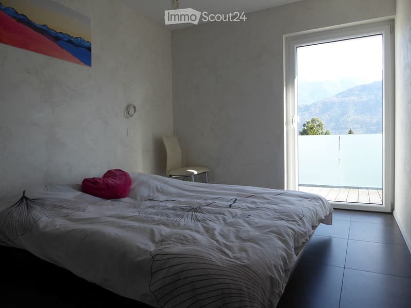 Modern bedroom with a double bed, white bedding, a red throw pillow, a white chair, and a small stool near a large window offering a scenic mountain view.