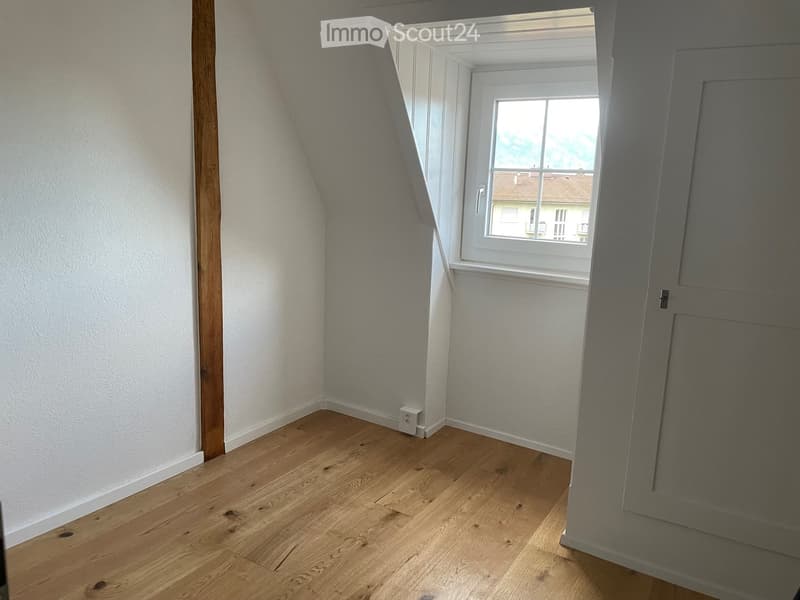 Empty room with wooden flooring, white walls, and a window showing a view of neighboring buildings.