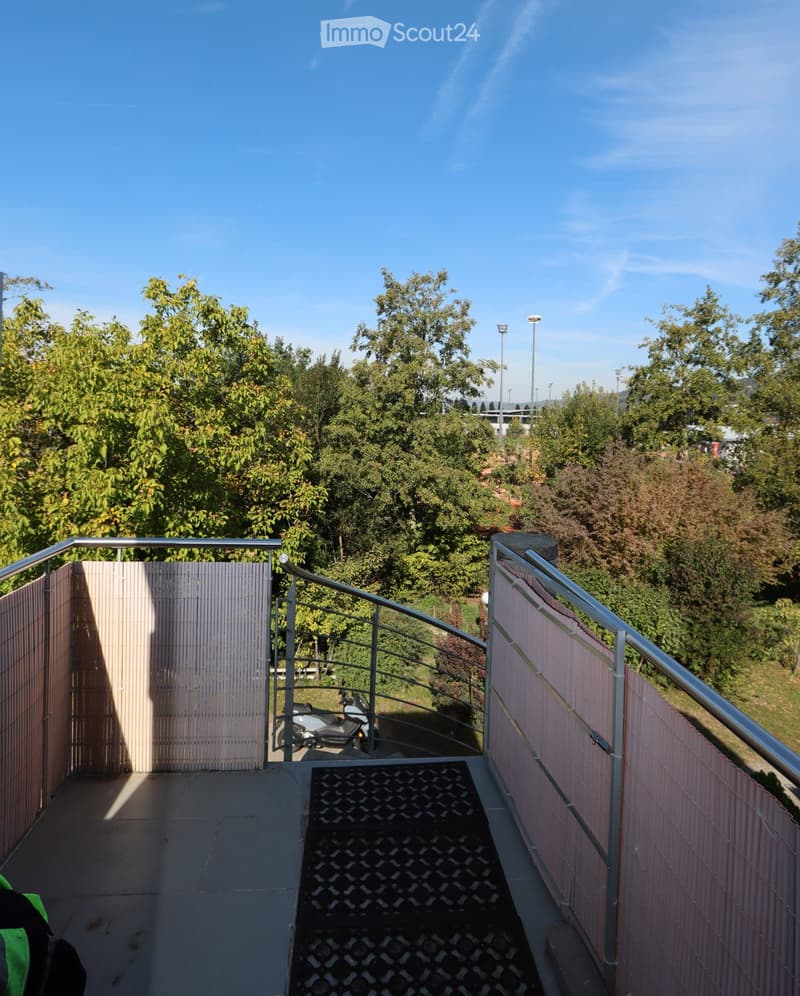 Balcony with metal railings, tiled floor, and a view of a green area with trees and a parked scooter.