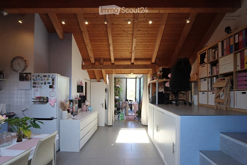 Open-plan living and dining area with exposed wooden beams, modern kitchen, dining table, office area, and built-in shelving.