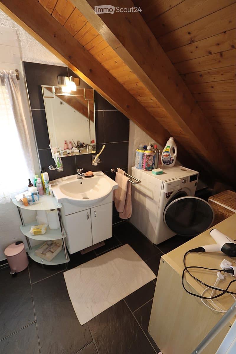 Bathroom with a sink, mirror, washing machine, and various toiletries. The room features dark tiled flooring and wooden slanted ceilings.