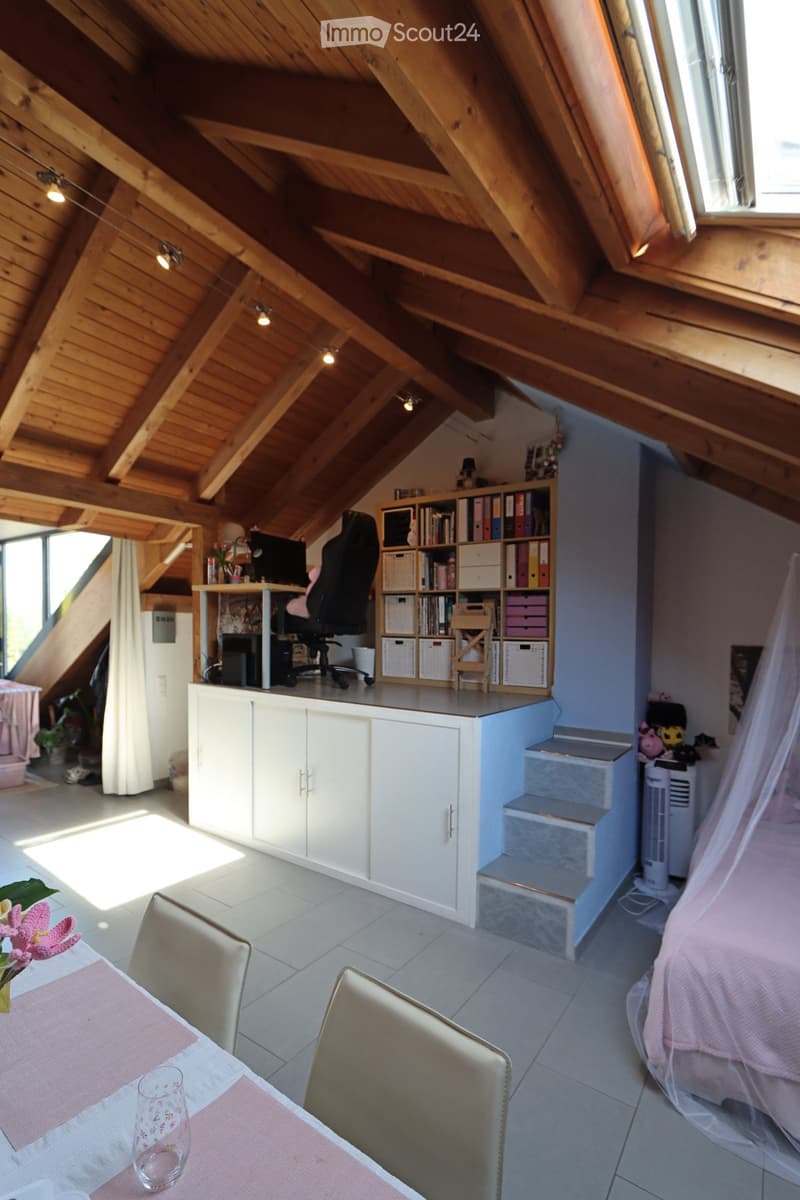 Loft-style room with wooden slanted ceilings, large windows, a bed with a pink cover, a desk with a computer, a bookshelf, a small table with a glass, and a fan.