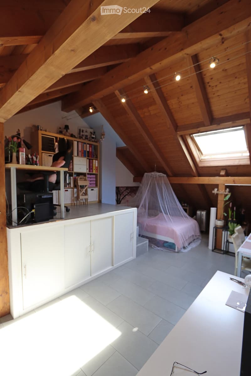 Spacious attic room with wooden beams, white tiled floor, and large skylight. Features a bed with a mosquito net, a desk with a laptop, a bookshelf, and a small seating area.
