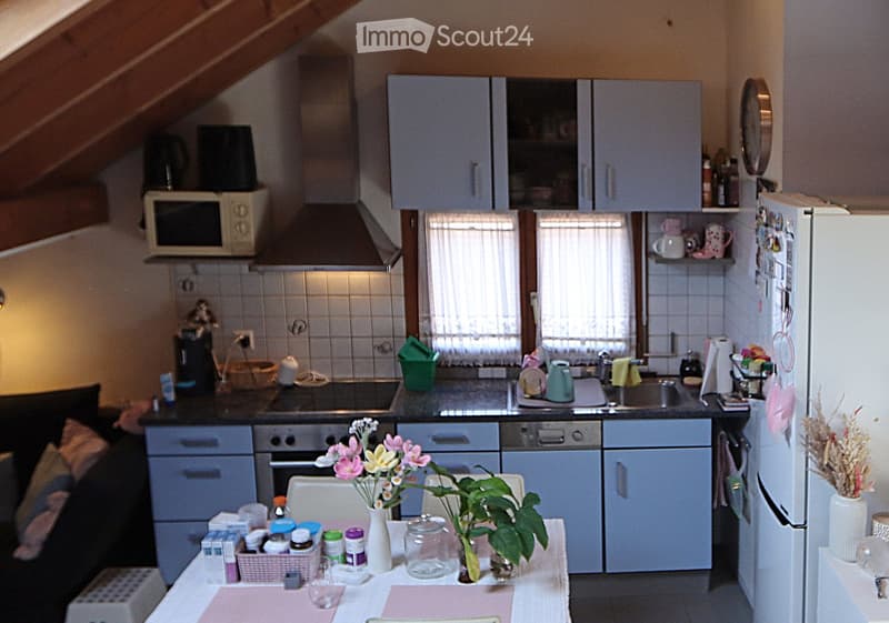 The image shows a cozy kitchen with light grey cabinets, a white refrigerator, and a microwave on a countertop. There is a dining area with a table and chairs, a vase with flowers, and a potted plant. The kitchen features a sink, stove, and various small