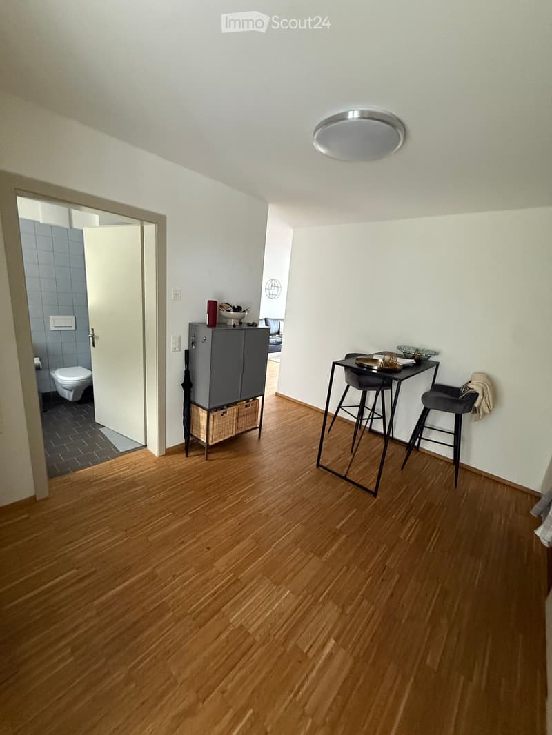 A small, modern room with wooden flooring, a round table with two stools, a grey cabinet with a basket underneath, and an open door leading to a bathroom.