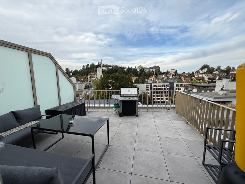 Modern rooftop terrace with a grey sofa, coffee table, barbecue, and storage box, overlooking a cityscape.