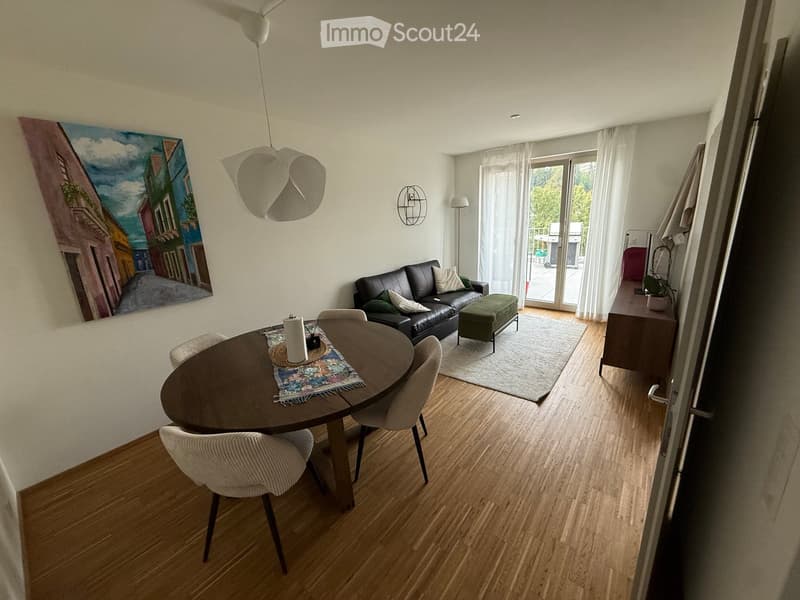 Open-plan living and dining area with wooden flooring, a black leather sofa, ottoman, round dining table with chairs, pendant light, wall art, side table with plant, and large window with white curtains leading to a balcony.