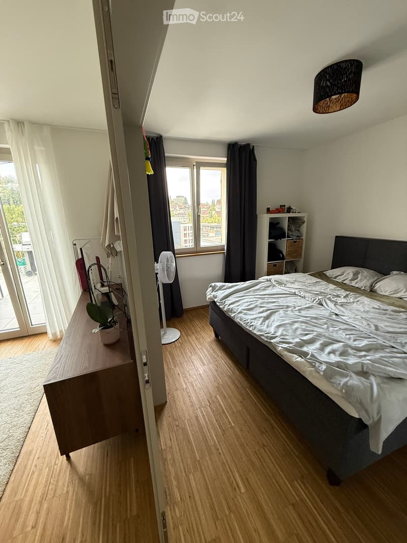 Modern bedroom with wooden flooring, large bed with white bedding, nightstand, fan, potted plant, and shelves.