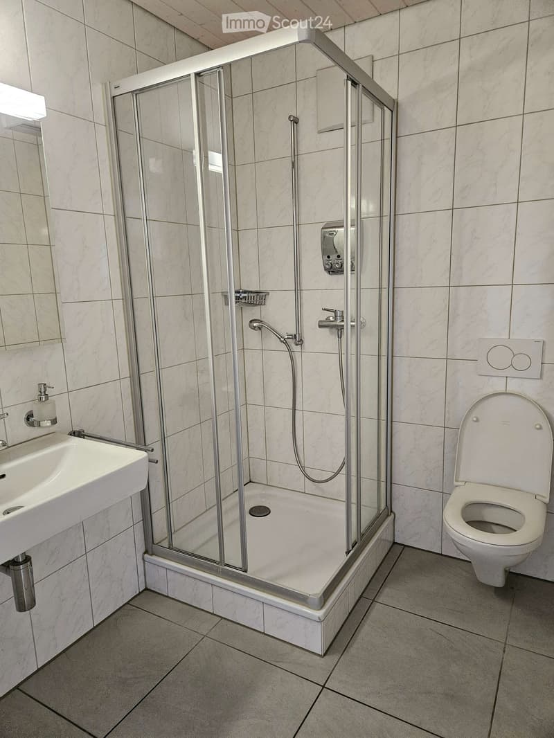 Modern bathroom with a white tiled shower enclosure, a sink with a mirror, and a toilet. The shower features a handheld sprayer and a built-in shelf. The walls and floor are tiled in a light marble pattern.