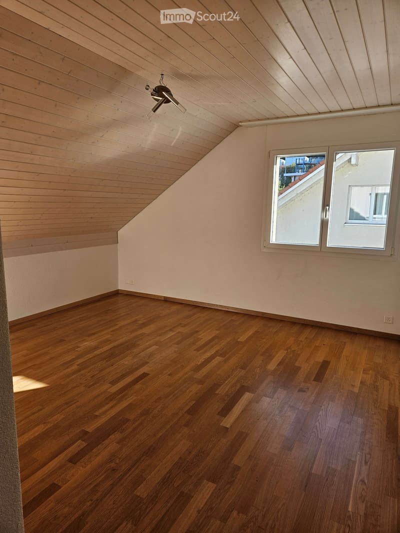 Empty room with wooden flooring, light-colored walls, and a slanted wooden ceiling. Features a ceiling fan and two windows with white frames.