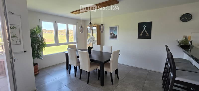 Bright dining room with a dark wooden dining table surrounded by white chairs, pendant lights hanging above, and a bar area with bar stools. Large windows offer a view of a green field.