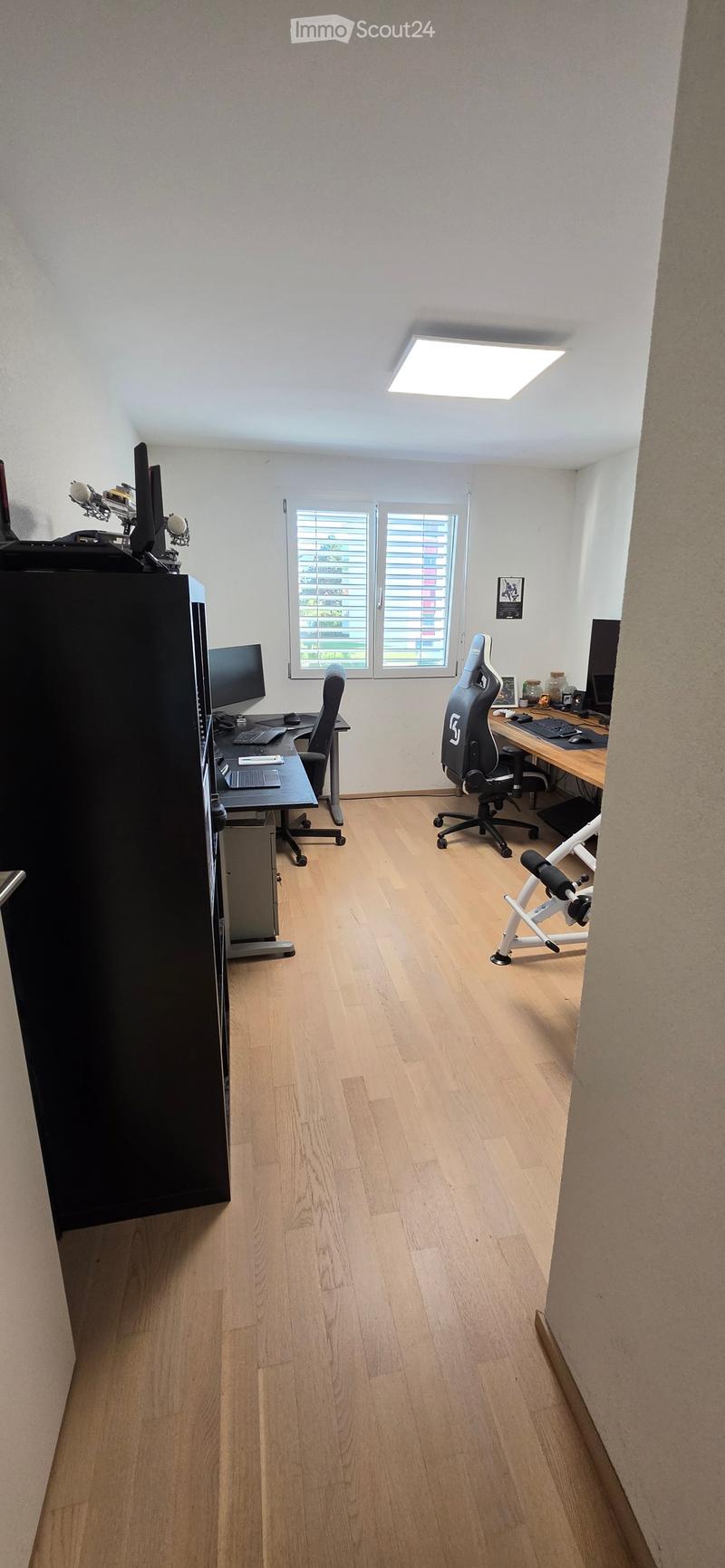 A well-lit room with wooden flooring, two desks with computers, chairs, and a fitness bike in the corner.