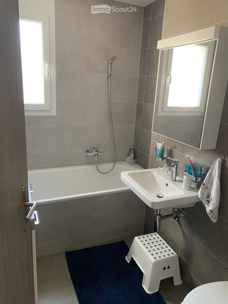 Modern bathroom with a bathtub, sink, mirror, and a small white stool. The walls are tiled in a neutral color, and the floor has a blue bath mat.