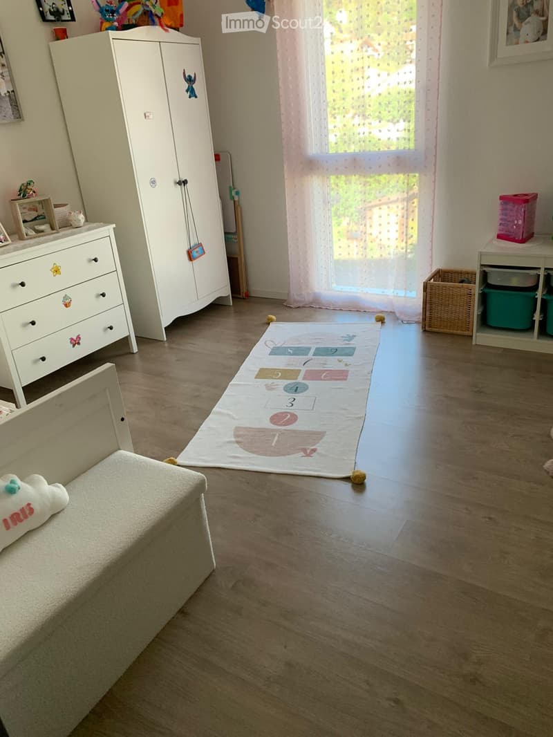 Child's room with wooden flooring, large window with sheer curtains, wardrobe, drawer unit, toy storage unit, play mat, and a small stool with a plush toy on it.