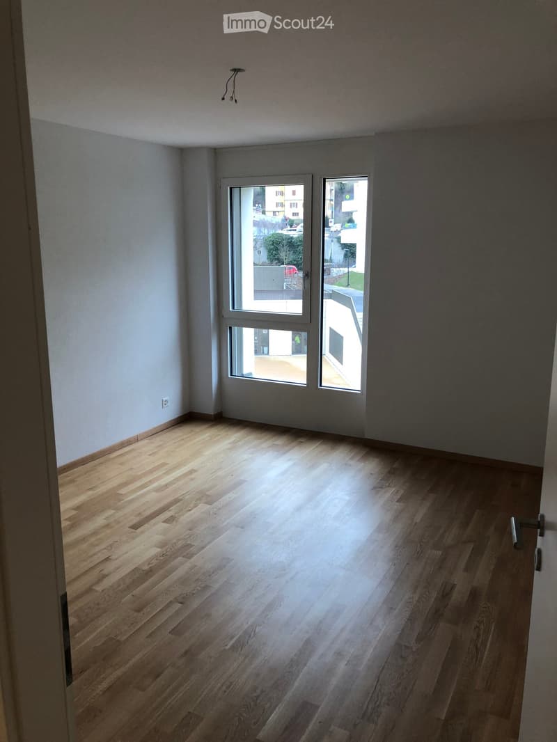 Empty room with light wooden flooring, white walls, and large windows offering a view of the street.