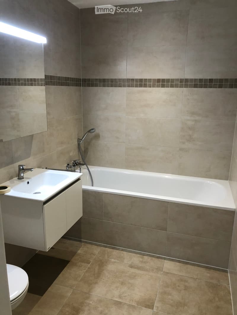 Modern bathroom with beige tiles, a white sink with a chrome faucet, a bathtub with a handheld shower, and a toilet. The walls are tiled in a combination of beige and dark brown, with a small mirror above the sink.