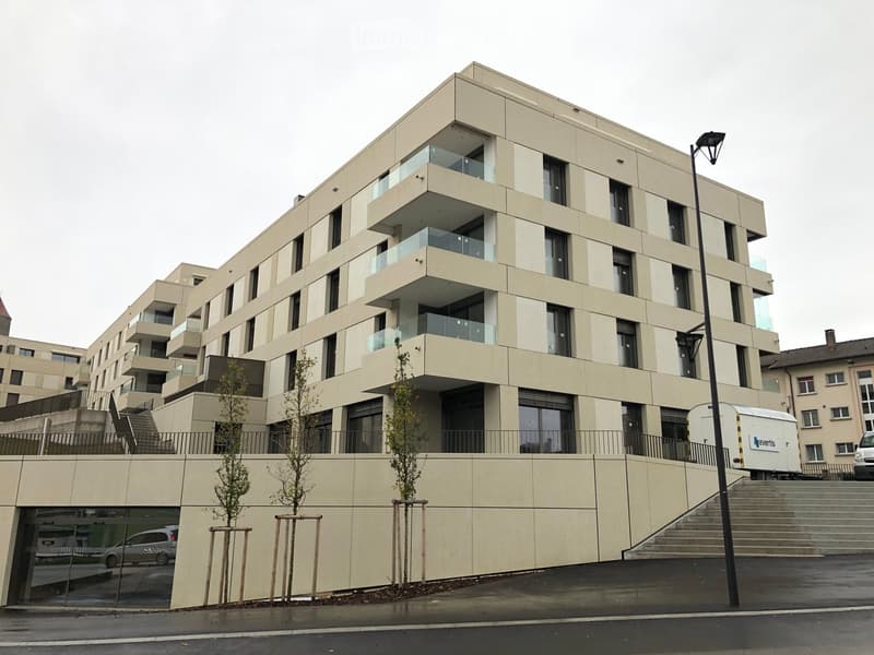 Modern multi-story residential building with beige facade, large windows, and balconies. Features a raised ground floor entrance and small landscaped area with young trees.