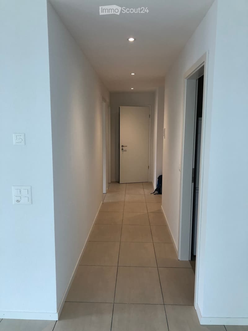 A clean, well-lit hallway with beige tiled flooring and white walls, featuring recessed ceiling lights and a closed door at the end.