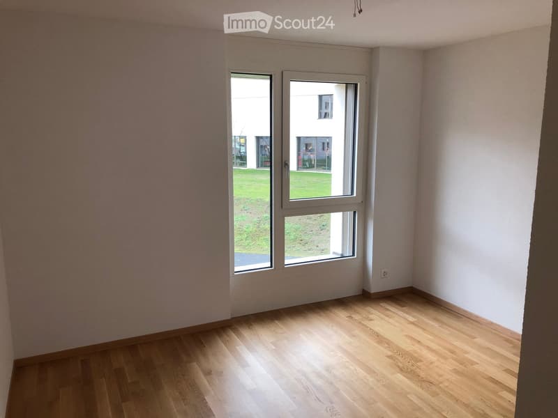 Empty room with light wooden flooring and white walls, featuring large windows with white frames that open to a view of a green lawn and neighboring building.