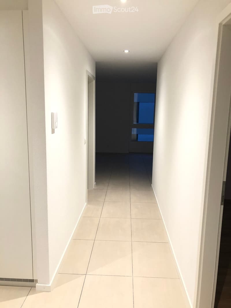 Clean, modern hallway with white tiled flooring and white walls, featuring recessed lighting and a door at the end.