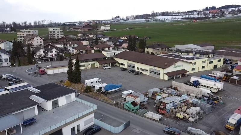 Aerial view of a commercial area with multiple buildings, parking lots, and various vehicles including cars, vans, and trailers.