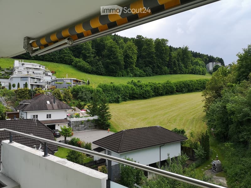 View from a balcony showing a green, hilly landscape with several houses, trees, and a well-maintained lawn.