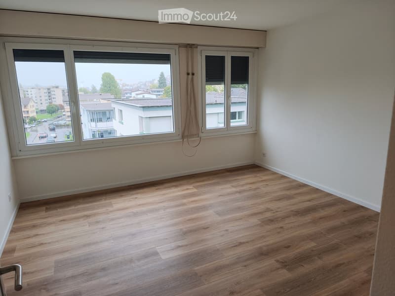 Empty room with wooden flooring and large windows offering a view of the street and surrounding buildings.