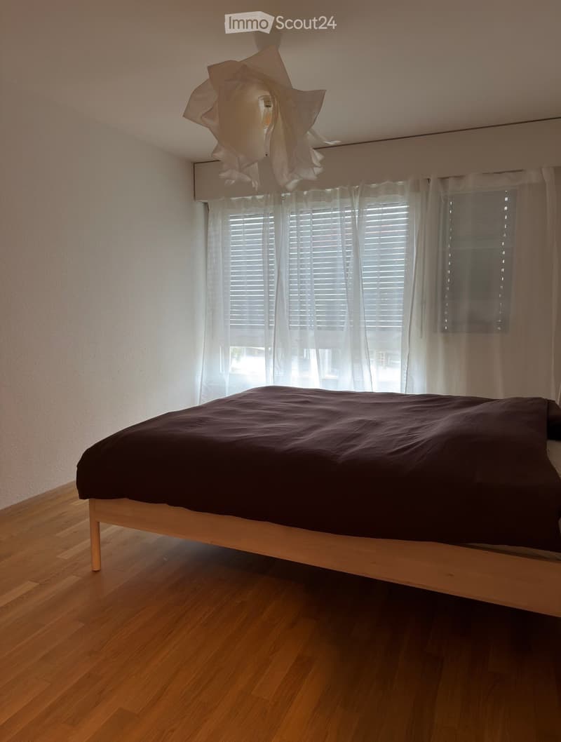 A modern bedroom with a wooden bed, dark bedding, large windows with white curtains and blinds, and a decorative hanging light fixture.