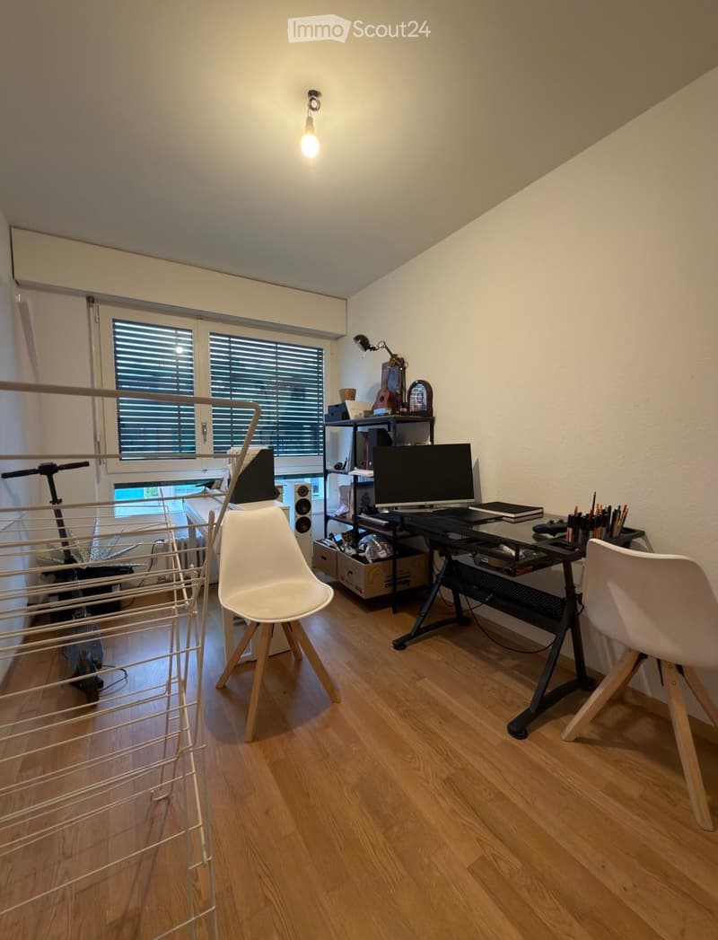 A modern, well-lit room with wooden flooring, a desk setup, two chairs, a guitar, and a balcony view.