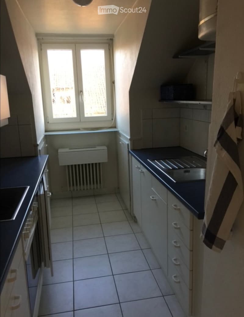 Empty kitchen with tiled floor, dark countertops, built-in cabinets, and a window with blinds. No appliances or furniture are present.