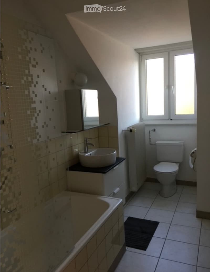 Modern bathroom with a bathtub, sink, toilet, and mirror. The space features tiled walls and a window providing natural light.