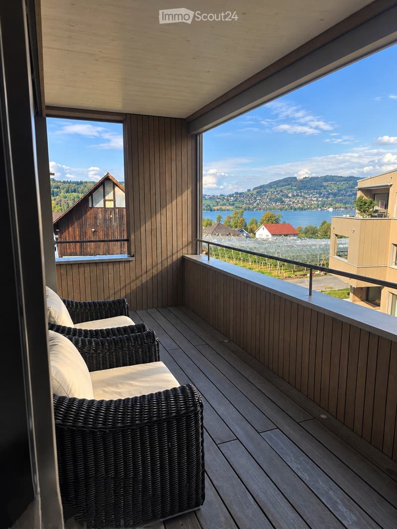 Balcony with wooden decking, wicker chairs, and scenic view of a lake and surrounding landscape.