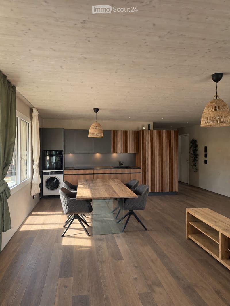Modern kitchen and dining area with wooden flooring, grey and wooden cabinets, a central dining table with chairs, pendant lights, and a washing machine.